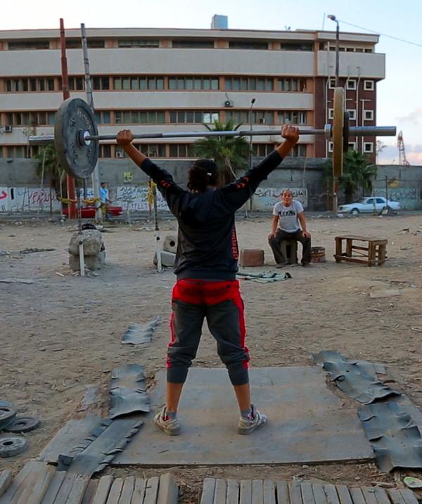 Lift Like a Girl, Szenenbild: Auf einem staubigen Platz stemmt eine Sportlerin vor den Augen eines älteren Mannes ein Gewicht.