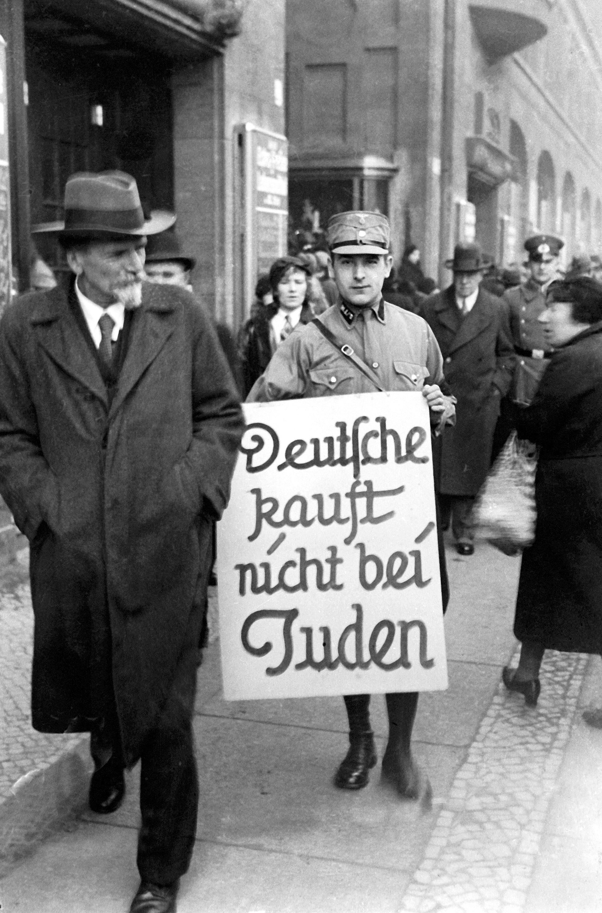 Berlin 1933 - Tagebuch einer Großstadt, Szenenbild: Das historische Schwarzweißbild zeigt einen Mann in SA-Uniform auf einem belebten Bürgersteig, der ein Schild mit der Aufschrift "Kauft nicht bei Juden" in den Händen hält