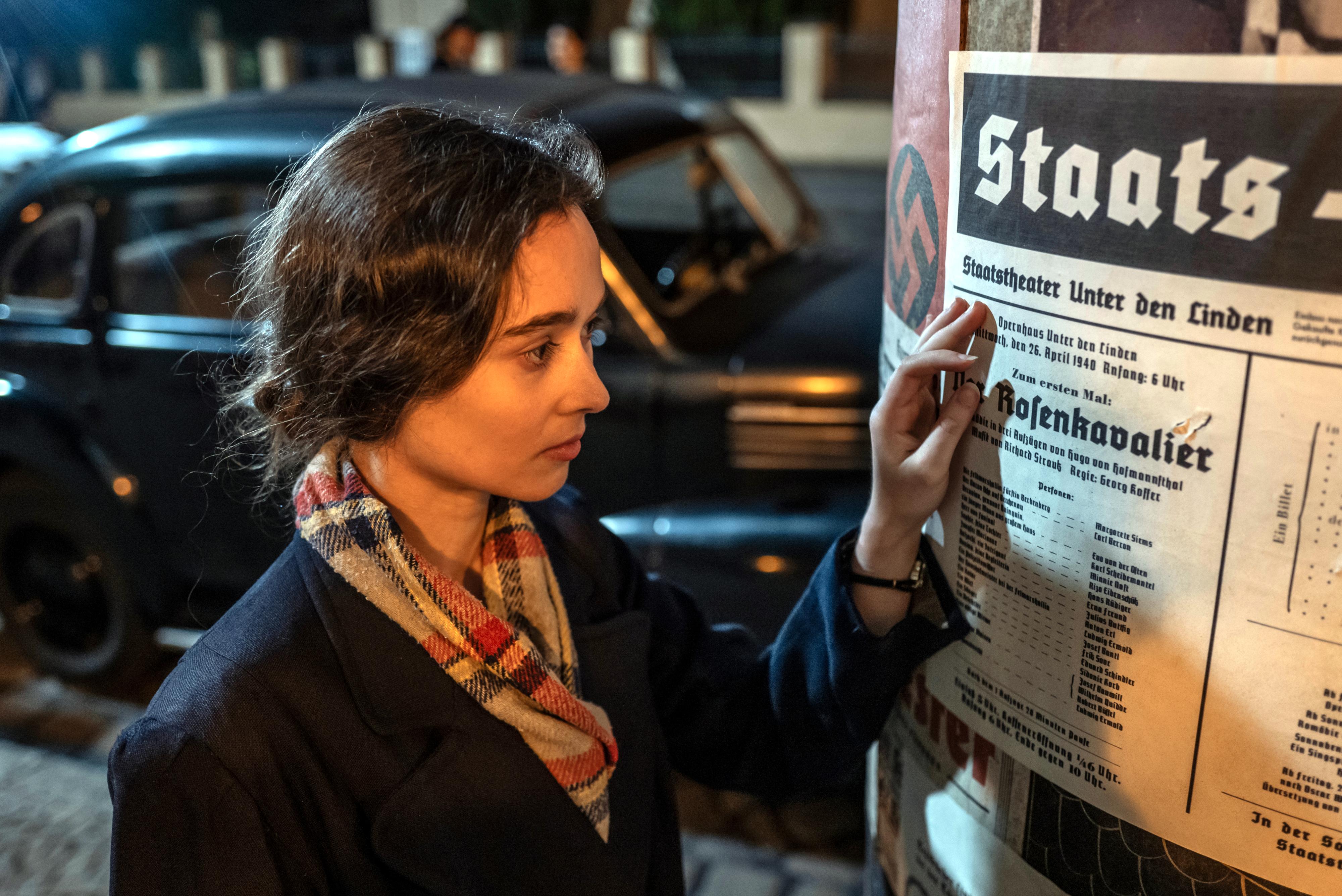 Ich bin! Margot Friedländer, Szenenbild: Eine junge Frau steht vor einer Lifaßsäule und ließt ein Werbeplakat für eine Theateraufführung