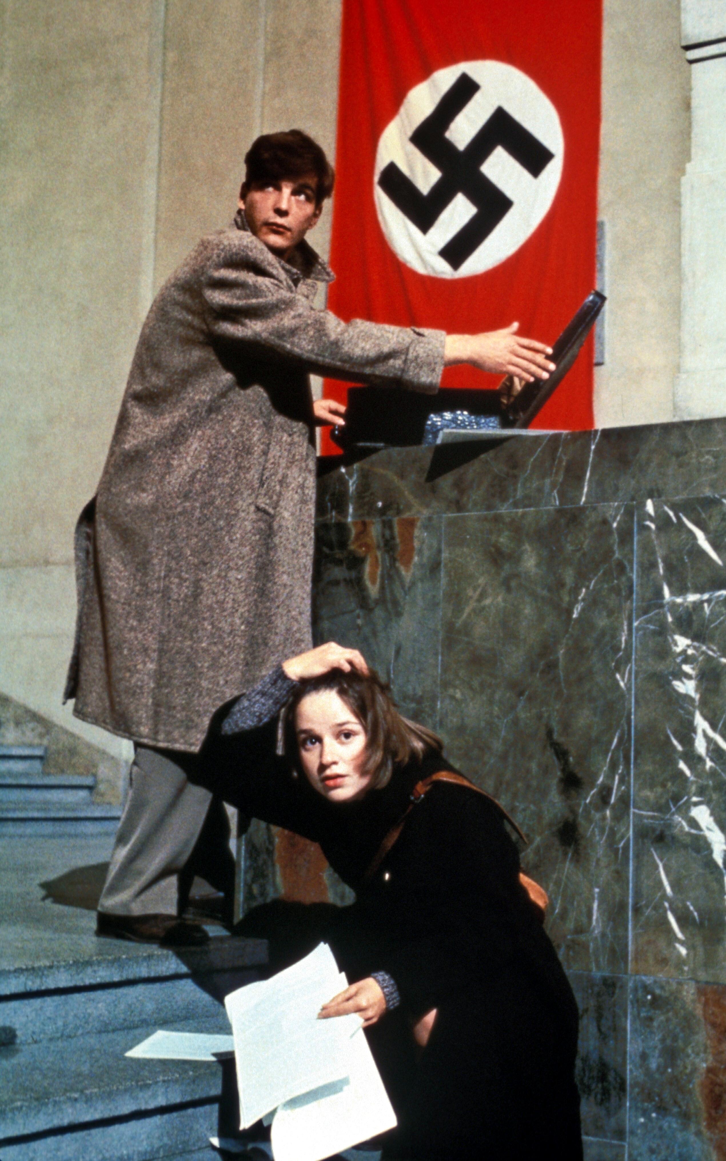 Szenenbild aus dem Drama "Die weiße Rose" (1982): Ein junger Mann hält steht auf einer Treppe und hält einen Koffer auf, der auf einen Vorsprung steht. Er schaut sich um. Neben ihm kniet auf der Treppe eine junge Frau. Sie hält Papierblätter in der H