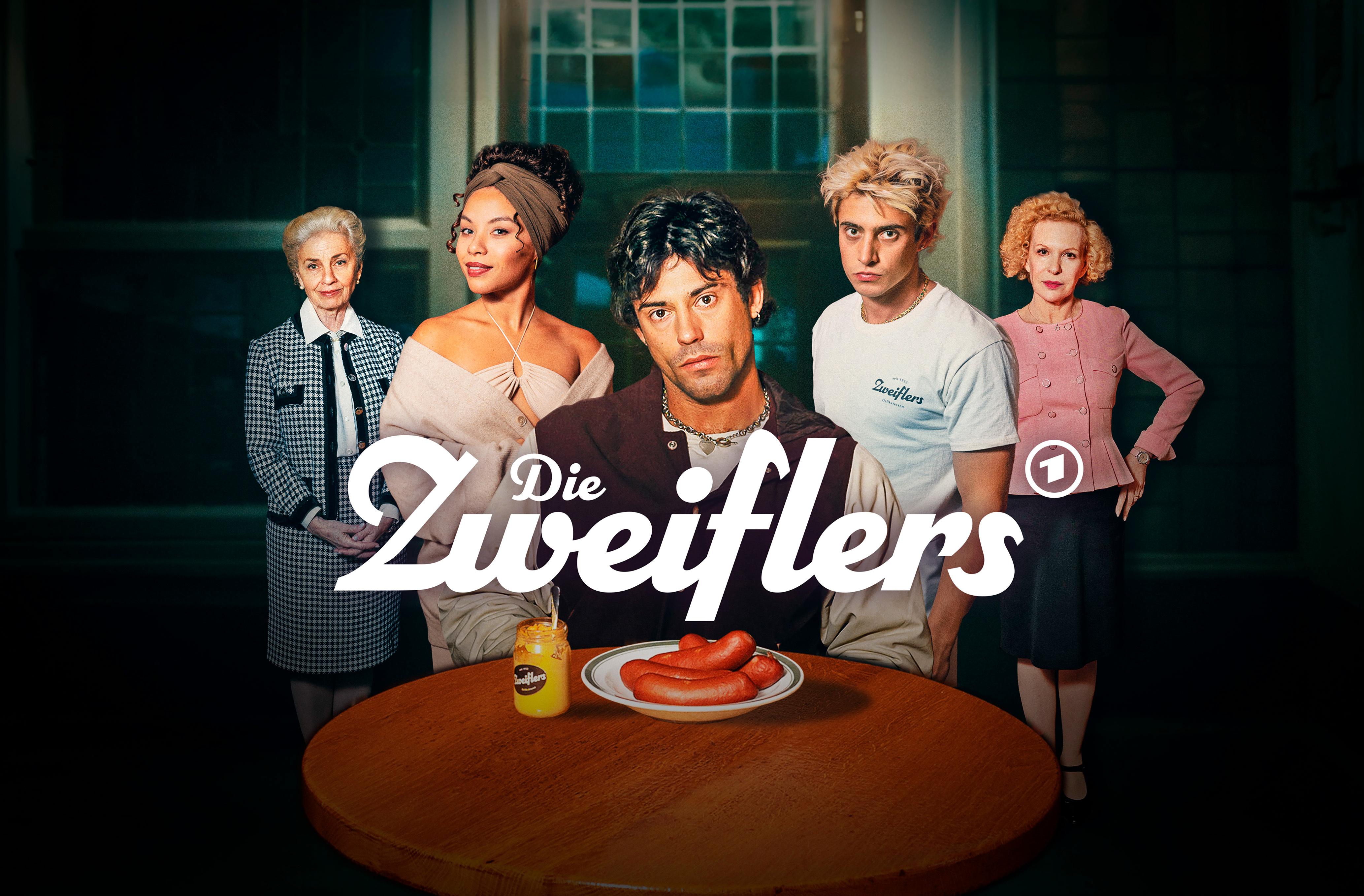 Key Visual zur Serie "Die Zweiflers": Ein junger, dunkelhaariger Mann steht vor einem runden Holztisch, auf dem ein Teller mit Würsten und ein Glas Senf steht. Der Mann ist gerahmt von drei Frauen unterschiedlichen Alters und einem weiteren Mann.