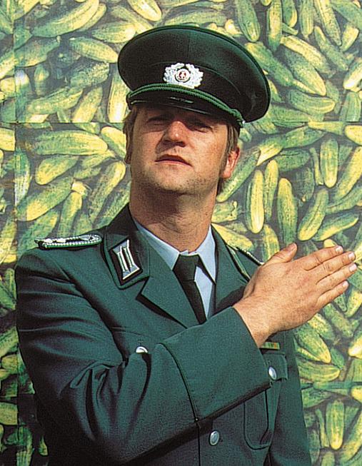 Szenenbild aus dem Film "Sonnenalle": Ein Mann in der Uniform der Volkspolizei steht vor einem Plakat, auf dem Gurken abgebildet sind, und blickt Richtung Kamera. Mit dem rechten Arm weist er nach rechts