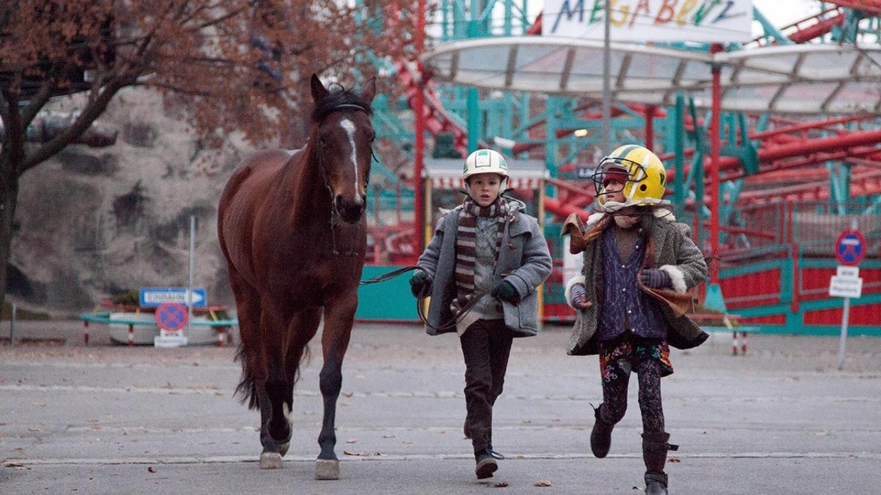 Szenenbild aus <filmtitel>Das Pferd auf dem Balkon</filmtitel>: Zwei Kinder laufen gemeinsam mit einem Pferd über einen Jahrmarkt.