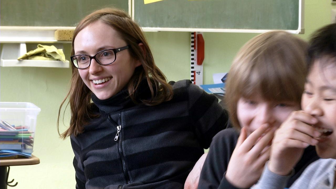 Szenenbild aus dem Dokumentarfilm <filmtitel>Zwischen den Stühlen </filmtitel>: Nahaufnahme einer jungen Lehrerin, die in einem Klassenzimmer neben zwei lachenden Kindern sitzt.n 