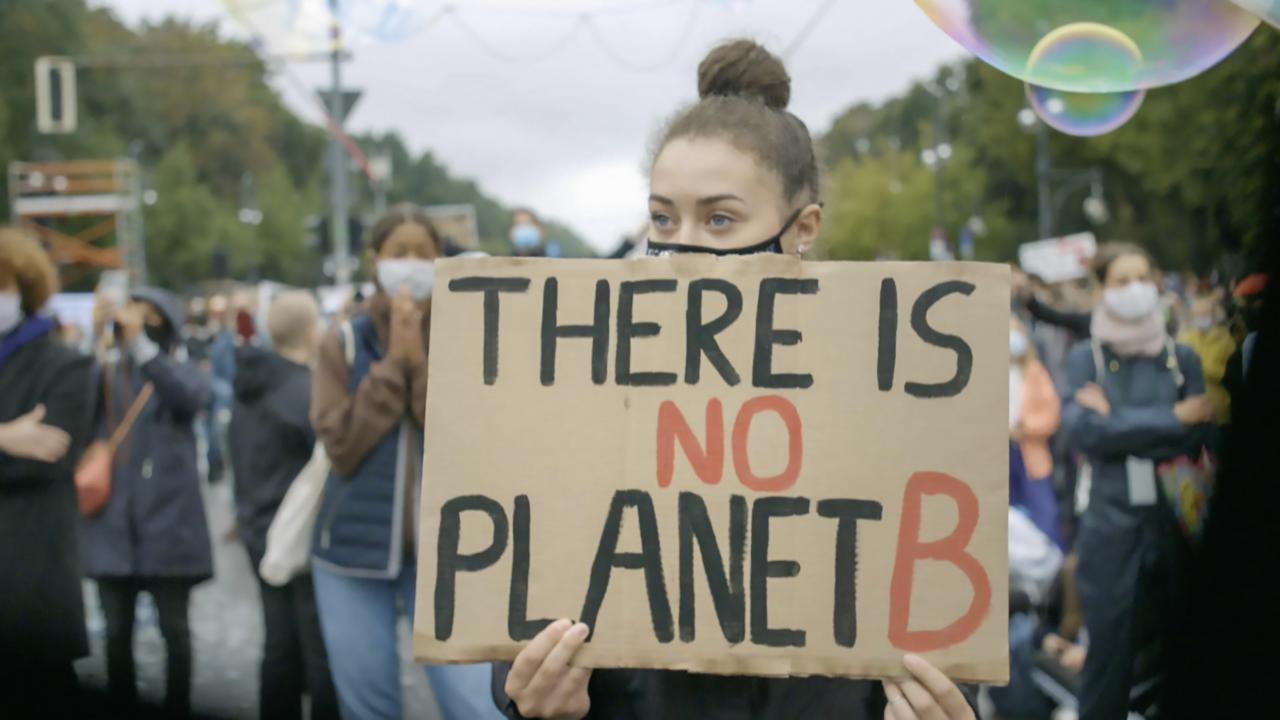 Eine junge Demonstrantin mit hochgesteckten Haaren hält ein Pappschild mit der Aufschrift "There is no planet b" in der Hand. Um ihr herum sind viele Menschen mit Masken. 