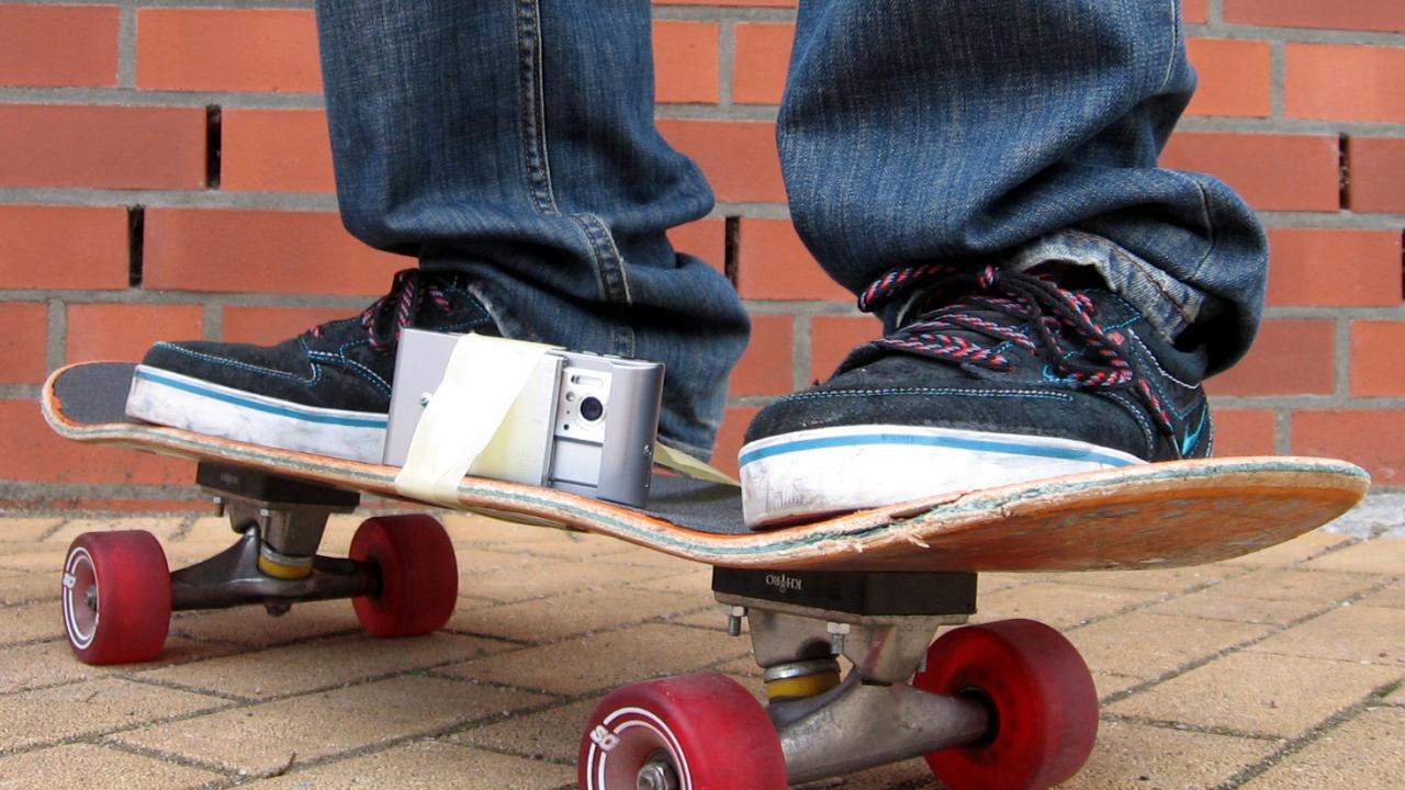 Nahaufnahme: Zwei Füße auf einem Skateboard, auf dem eine kleine Filmkamera festgeklebt ist.