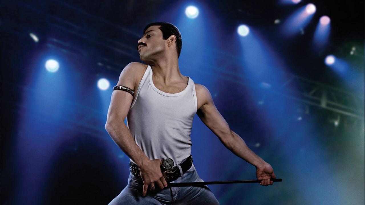 Der Schauspieler Rami Malek als Freddie Mercury, Sänger von der Band Queen, steht unterm Rampenlicht der Bühne. Er trägt ein weißes T-shirt, eine Jeans und ein Armband. Er hält eine Mikrophonestange mit beiden Händen und guck zur Seite. 