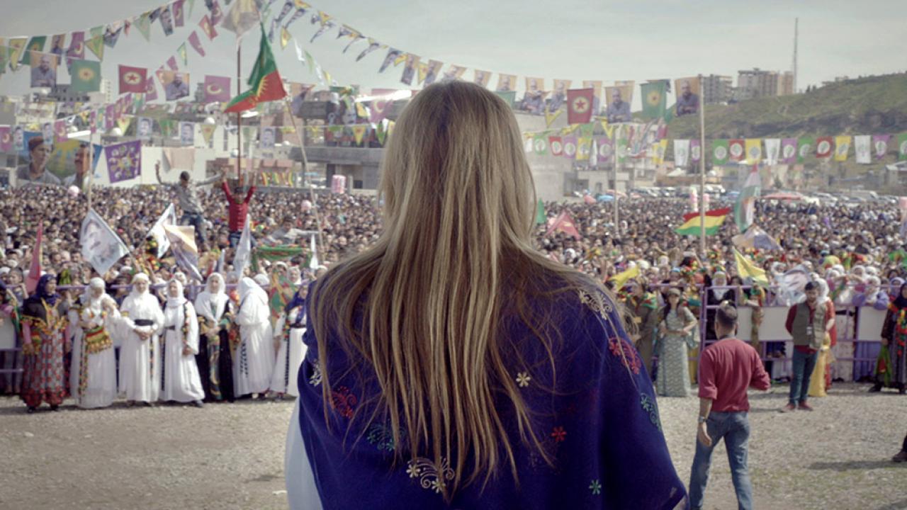 Nahaufnahme einer Frau mit langen blonden Haare. Sie steht mit den Rücken zur Kamera, vor einer Menge von Menschen, die kurdische Flaggen wehen. 