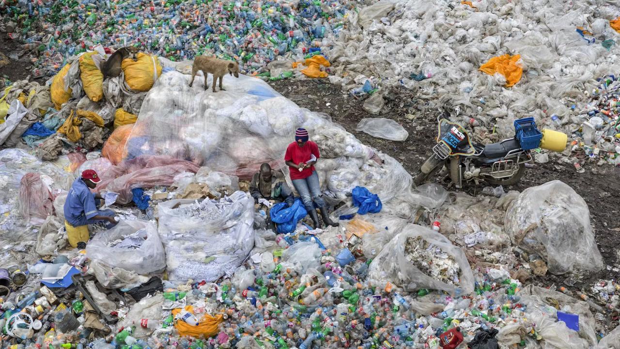 Totale einer Plastikmüllhalde mit Müllsammlern in der Nähe von Nairobi. In der Mitte lehnen sich zwei Menschen an den Müllberg, Links sammelt einere andere Figur etwas. Auf dem Berg steht ein Hund. 