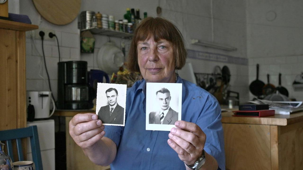 Eine ältere Dame im blauen Hemd zeigt dem Betrachter zwei Schwarz-Weiß-Fotografien von jungen Männer im Anzug. Sie sitzt in einer Küche. 