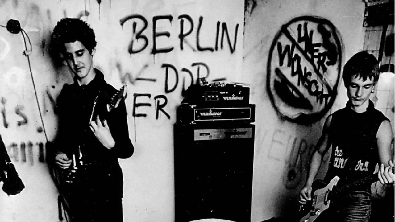 In schwarz-weiß: zwei junge Männer in punk-Kleidung spielen Guitare vor einer Wand voller Grafitti. 