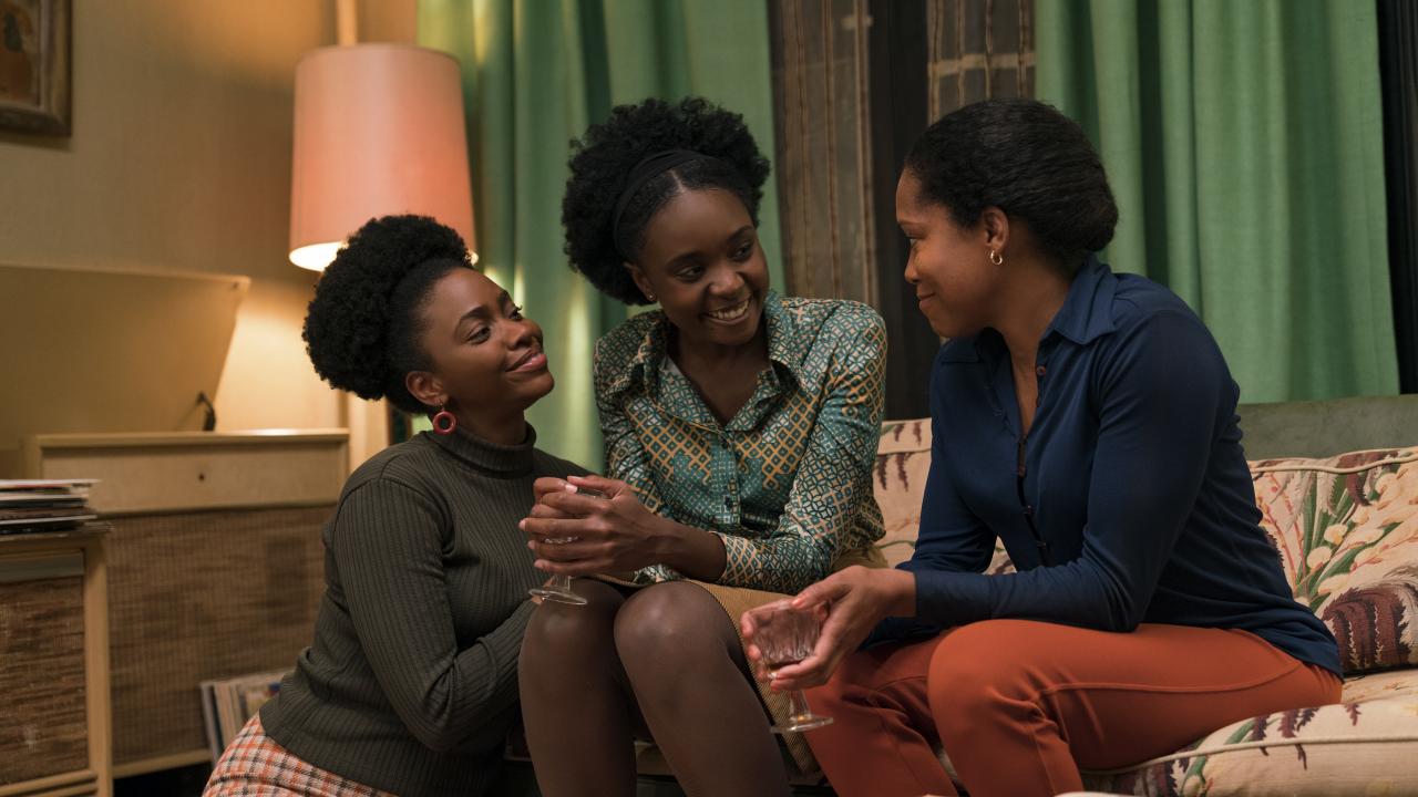 Zwei afroamerikanische Frauen sitzen auf einem Sofa, eine weitere sitzt auf dem Boden. Sie lächeln sich an.