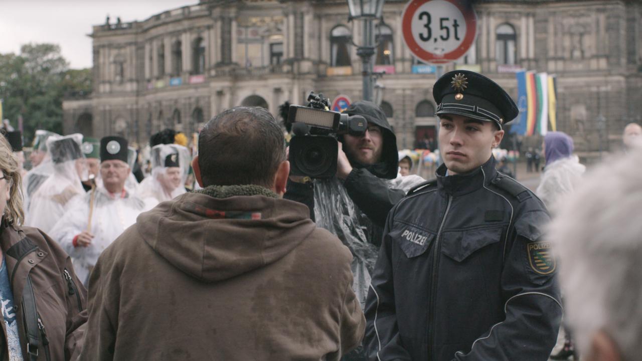 Eine Pegida-Demonstration in Dresden: Von hinten ist ein Mann zu sehen, der vor einem Medienvertreter mit Kamera steht, zwischen ihnen ein junger Polizist.