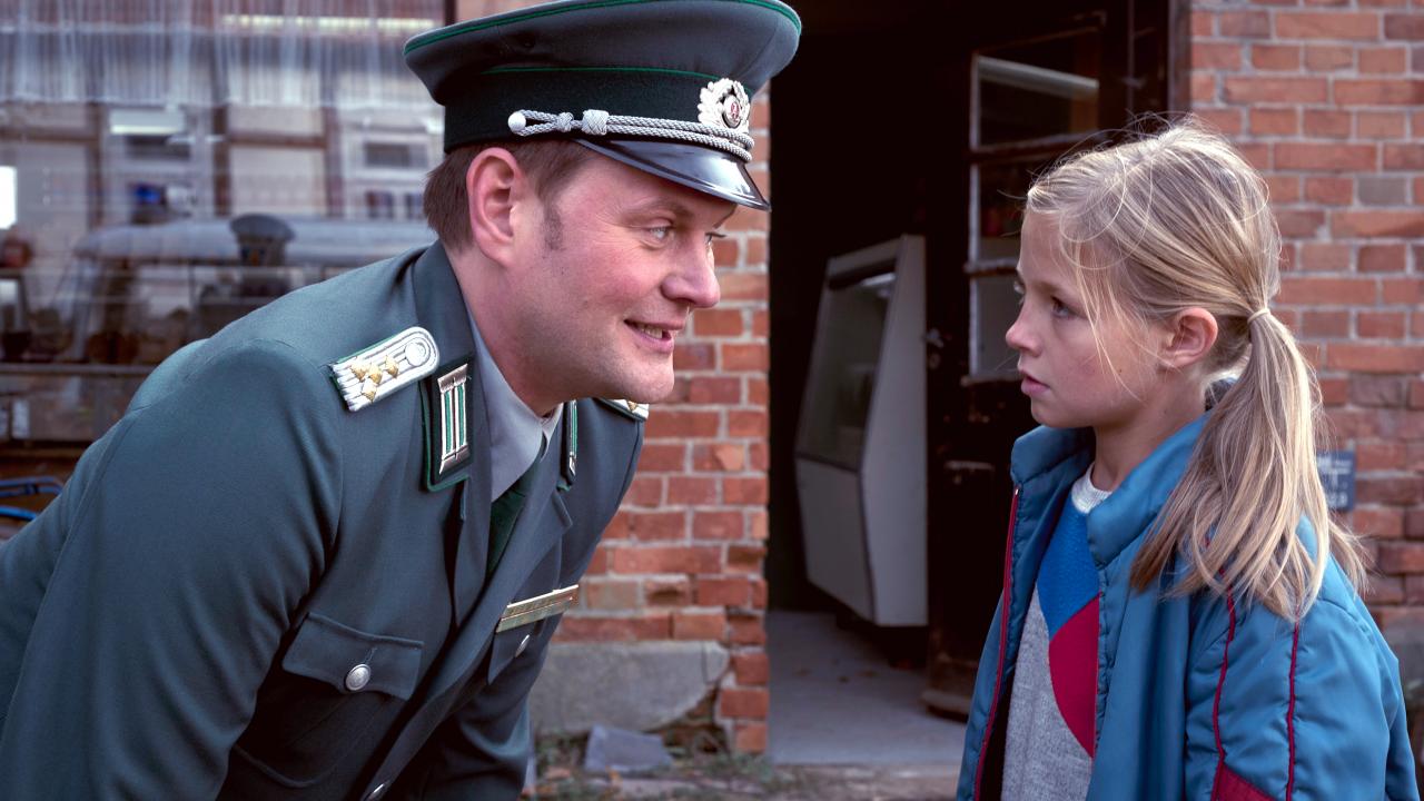 Szenenbild aus dem Kinderfilm "Sputnik": Ein DDR-Volkspolizist beugt sich zu einem Mädchen mit blonden Zöpfen herunter und spricht mit ihm.