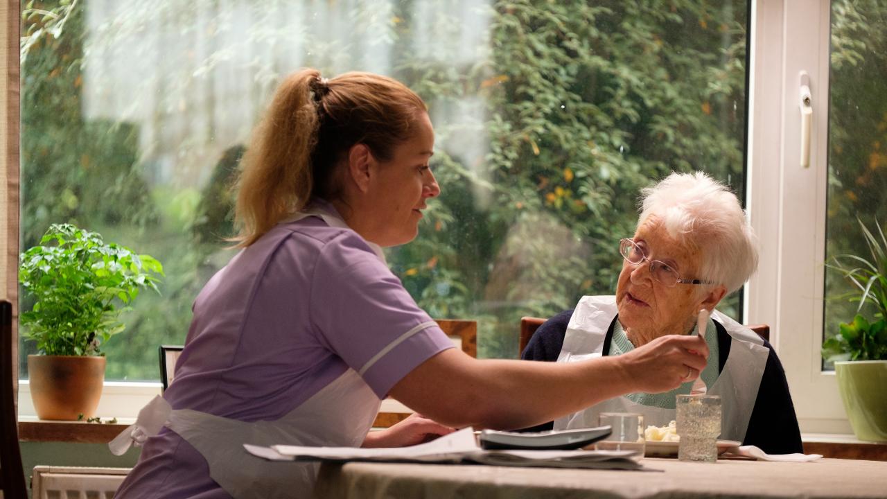 Eine Frau sitzt an einem Tisch und füttert eine alte Dame.