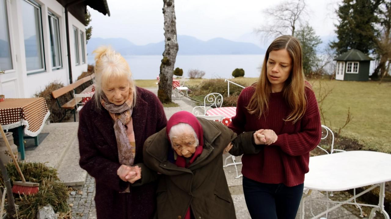 Szenenbild aus dem Dokumentarfilm "Walchensee Forever": Eine alte Dame wird beim Gehen von ihrer Tochter und ihrer Enkeltochter gestützt.