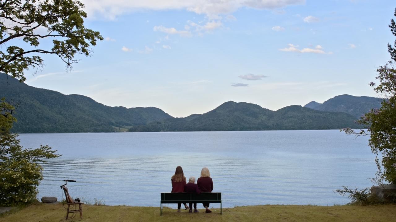 Szenenbild aus dem Dokumentarfilm "Walchensee Forever": Panoramablick auf den Walchensee und Alpen. In der Mitte des Bildes steht eine Bank, auf der in der Rückenansicht drei Frauen sitzen und auf den See blicken.