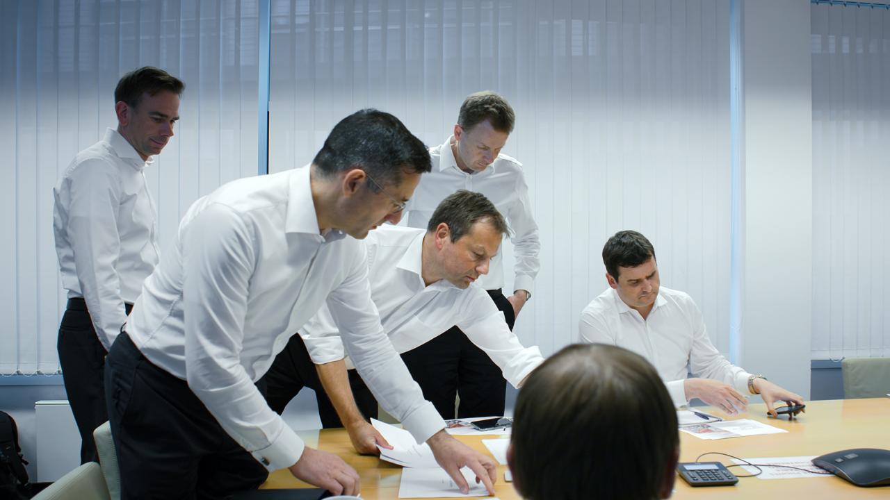 Mehrere Männer in Businesslook umringen  einen Bürotisch.