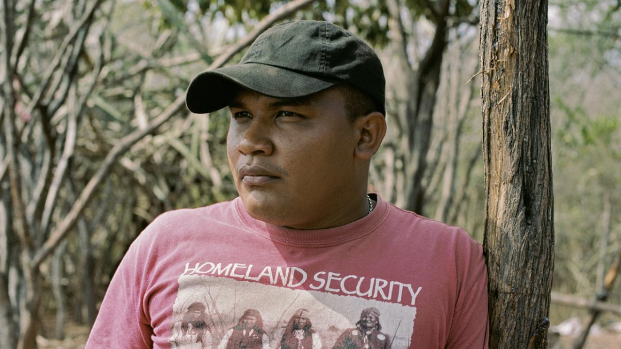 Mann mit schwarzer Kappe und rotem T-Shirt, "Homeland Security"-Aufdruck, steht im Wald und lehnt an einem Baumstamm.