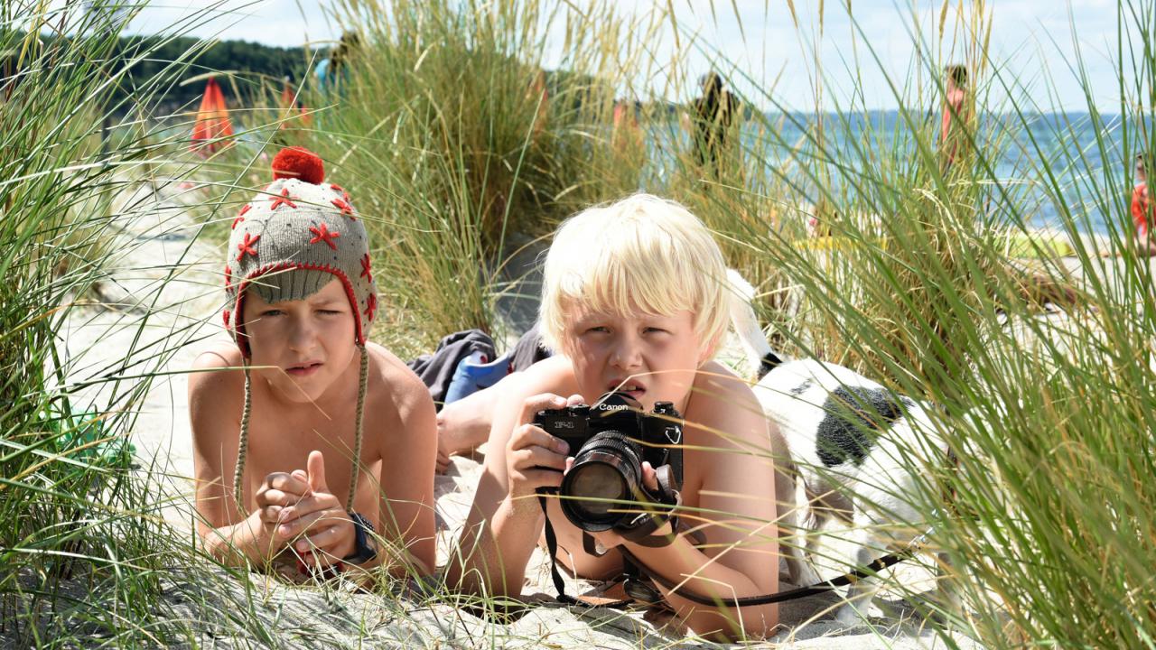 Zwei Jungen liegen am Strand im Gras, einer trägt eine Mütze, der andere hält eine Kamera. Im Hintergrund sind Meer und Sonnenschirme.