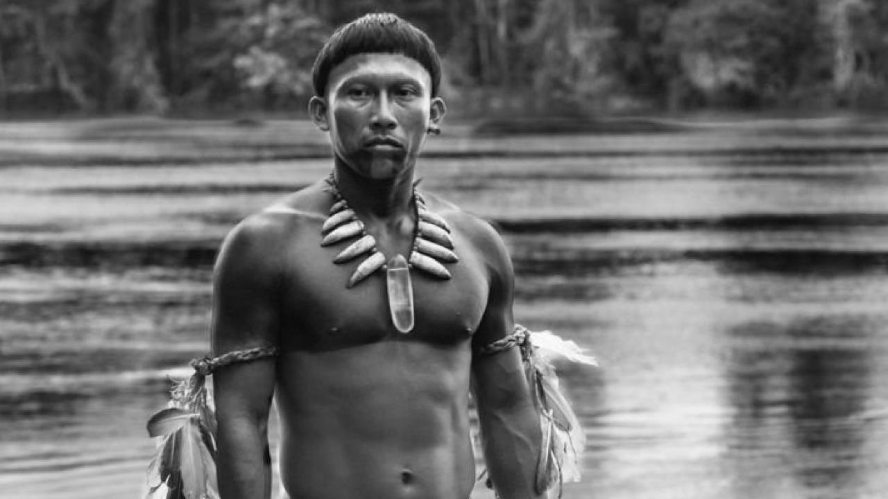 Indigener Mann am Fluss, mit traditionellen Schmuck und Kleidung. Schwarz-Weiß-Foto mit Wald im Hintergrund.