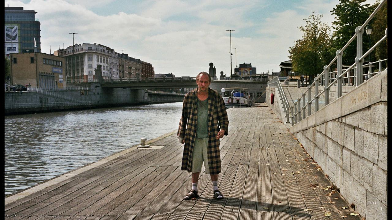 Älterer Mann in kariertem Mantel und Sandalen steht auf einem Holzsteg am Flussufer, Stadtgebäude und Brücke im Hintergrund.