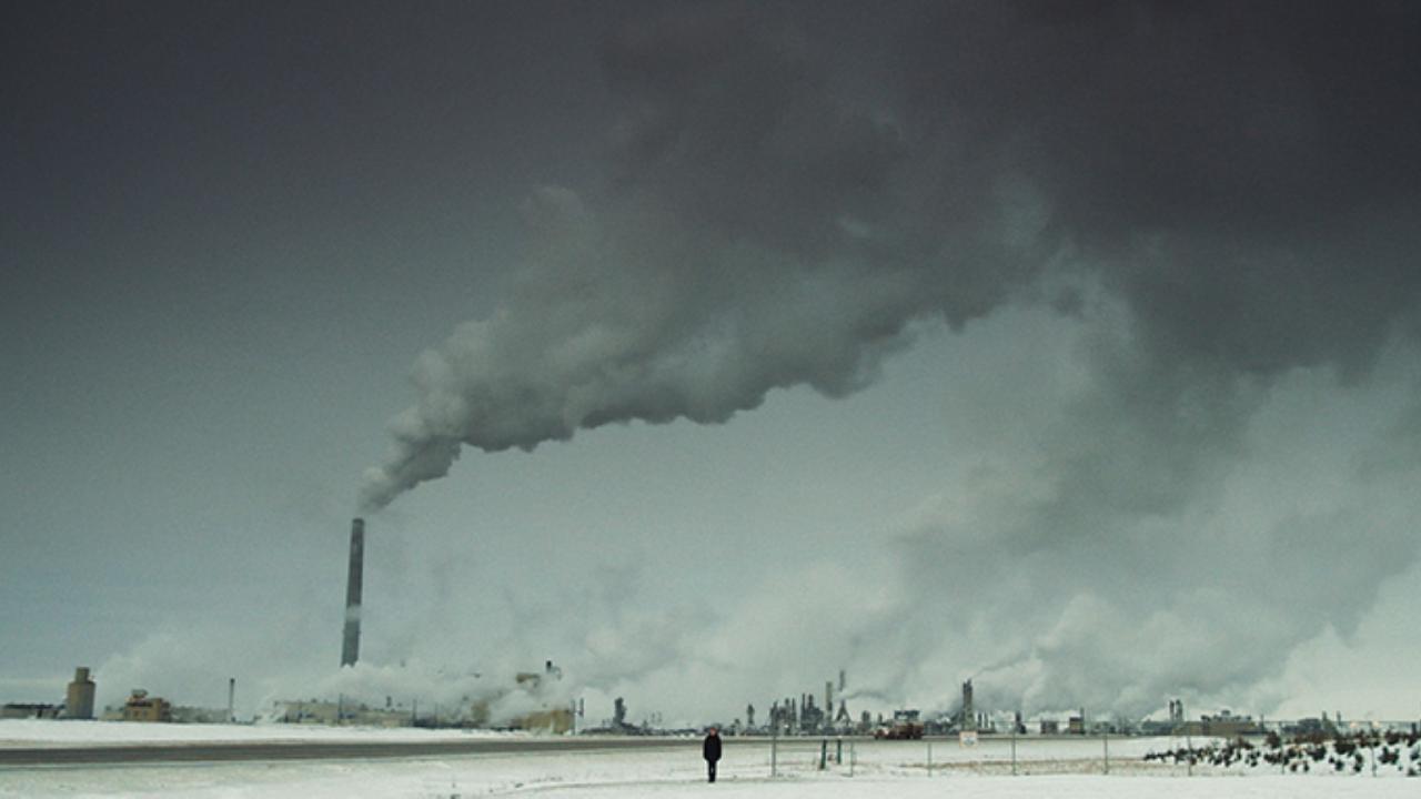 Totale einer winterlichen Landschaft. Im Hintergrund steht eine Industrienanalage mit rauchenden Schloten. Im Vordergrund steht als schwarze Silhouette eine Frau.