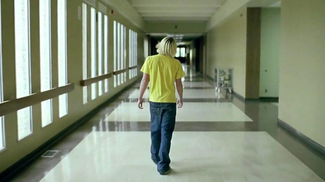 Junge mit blondem Haar und gelbem T-Shirt geht durch einen leeren Flur mit großen Fenstern.