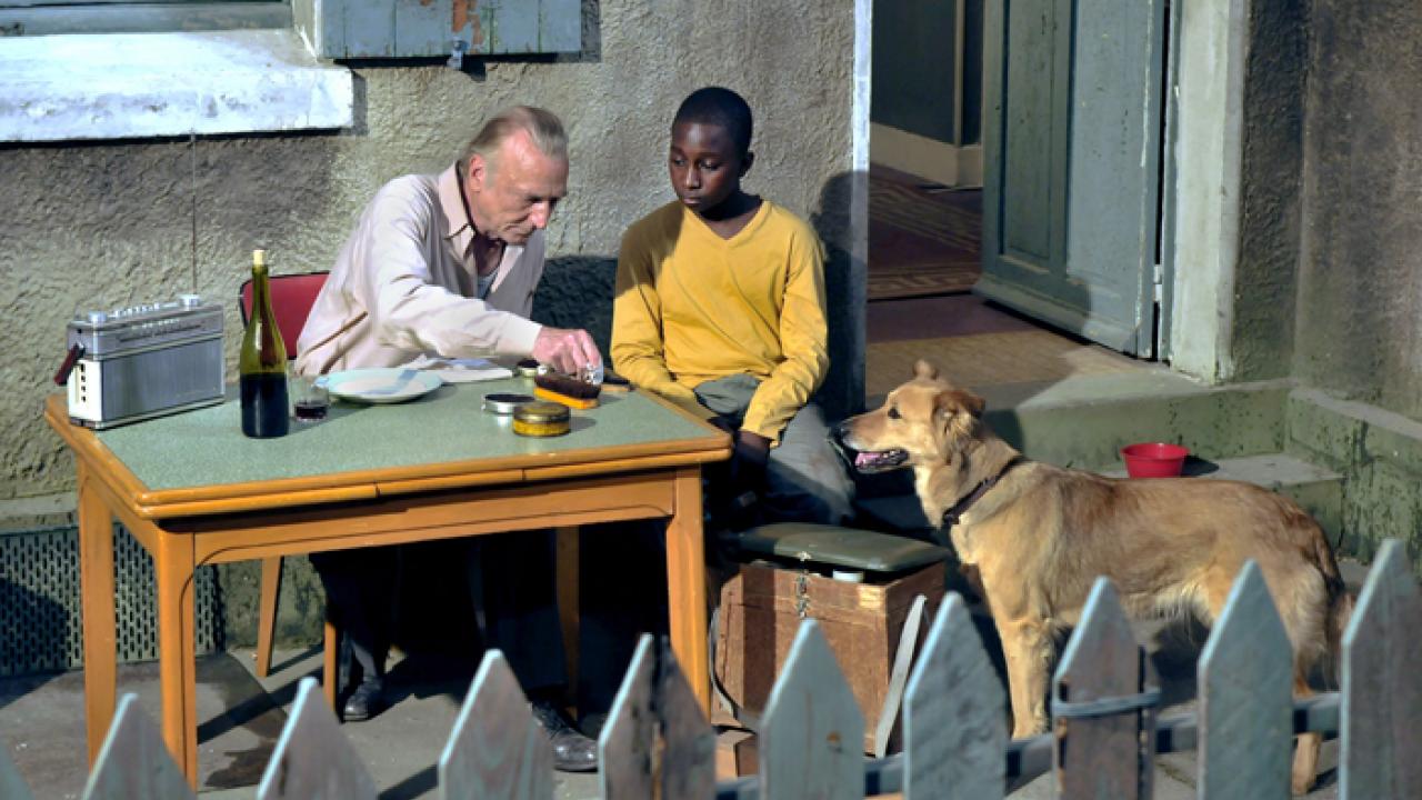 Älterer Mann und Junge sitzen draußen an einem Tisch, Hund steht daneben. Flasche und Radio auf dem Tisch.