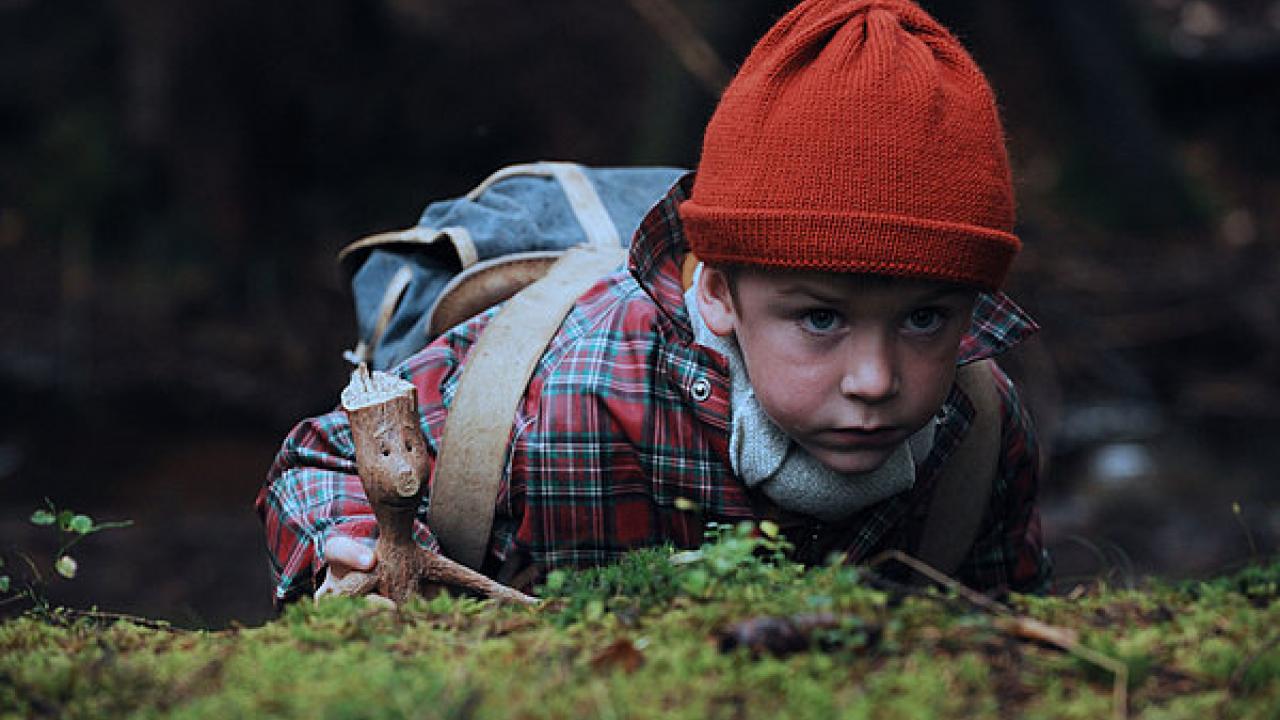 Kind mit roter Mütze und kariertem Hemd liegt im Wald, hält eine Holzfigur in der Hand.