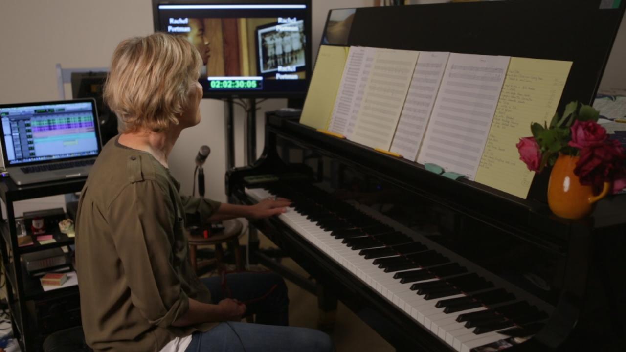 Blonde Frau sitzt am Klavier, Notenblätter aufgestellt, Monitor und Laptop im Hintergrund, Vase mit Blumen rechts.