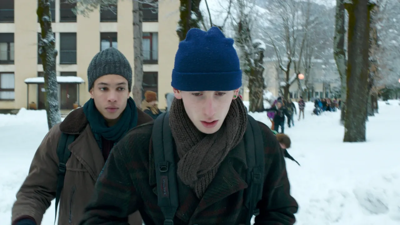 Zwei junge Männer in Winterkleidung gehen durch eine schneebedeckte Straße, umgeben von Gebäuden und Menschen.