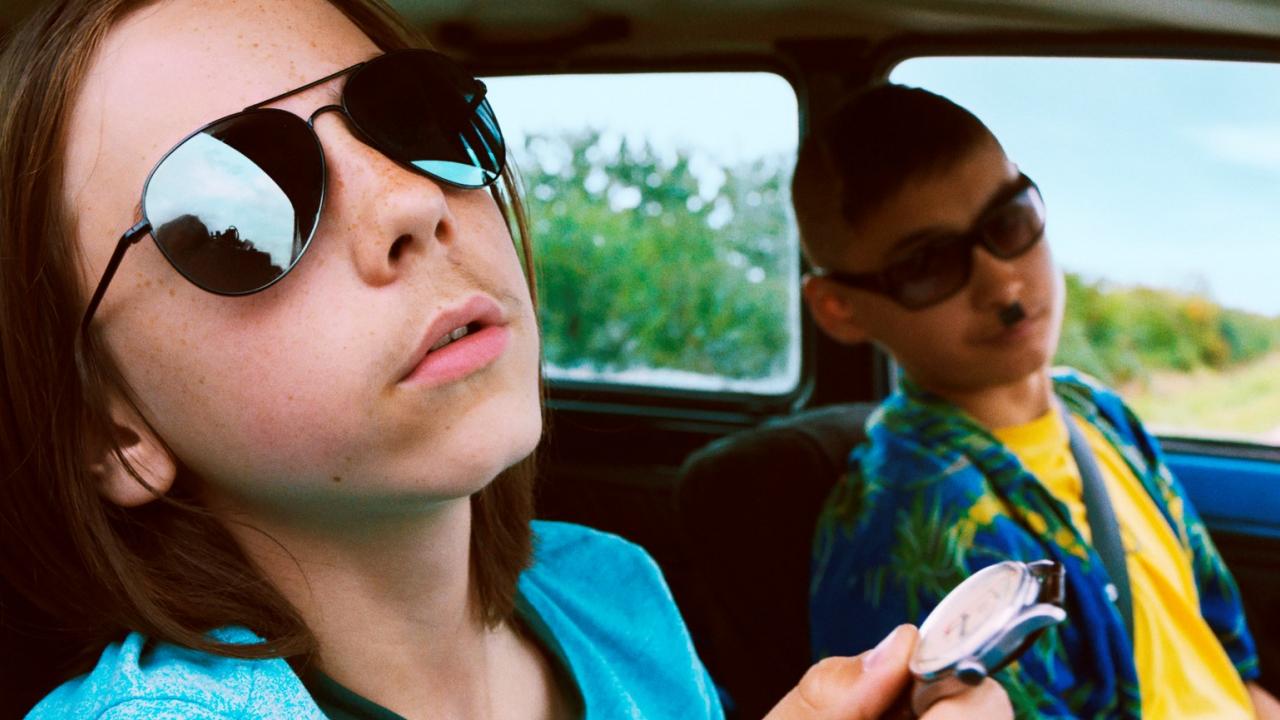 Zwei Jungen mit Sonnenbrillen im Auto, einer hält eine Uhr. Helle Kleidung und sommerliche Stimmung.