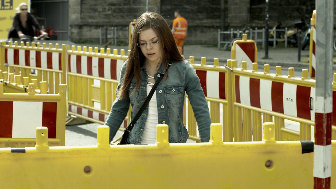 See You, Szenenbild: Eine junge Frau bahnt sich einen Weg durch eine abgesperrte Straße.