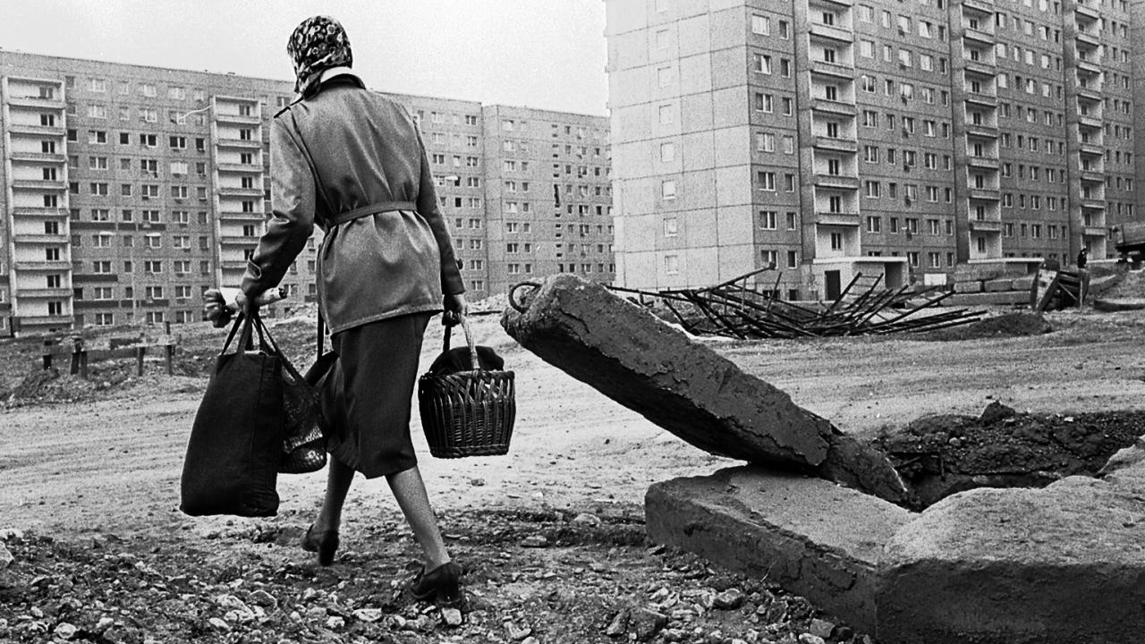 Schwarz-weißes Szenenbild aus dem Dokumentarfilm DIE UNBEUGSAMEN 2 - GUTEN MORGEN, IHR SCHÖNEN: Frau mit Einkaufstaschen geht über Trümmer in einer Neubausiedlung in Karl-Marx-Stadt (1978).