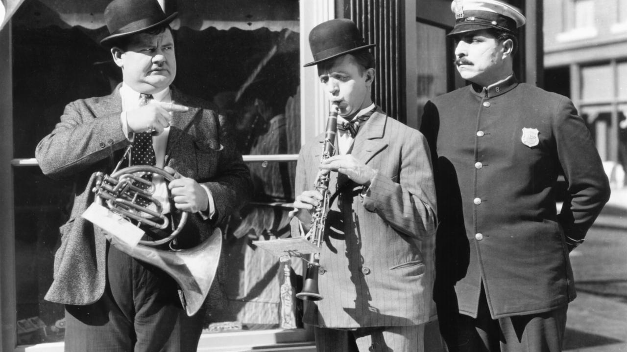 Szenenbild aus der Komödie LAUREL & HARDY: DER BELEIDIGTE BLÄSER: Der Komiker Stan Laurel, neben dem ein Polizist steht,  spielt auf einer Klarinette. Neben ihm steht Oliver Hardy und zeigt mit dem Finger auf ihn.