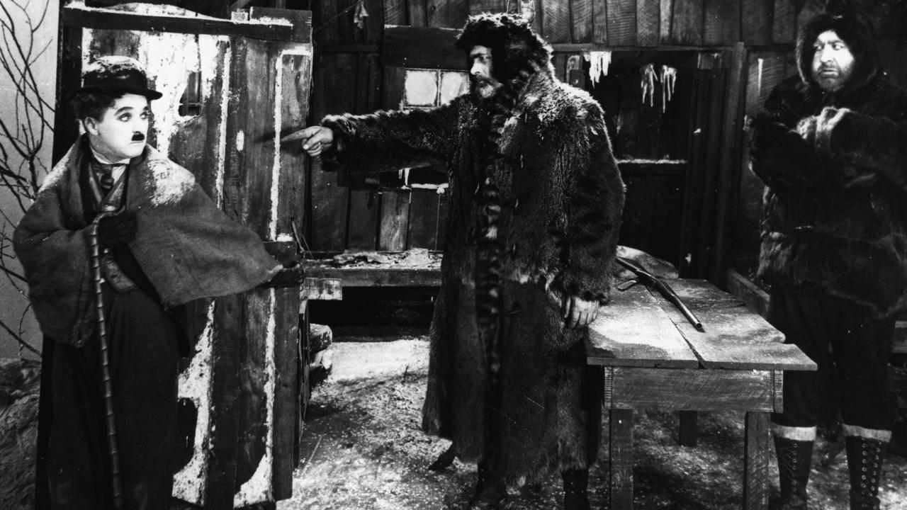 Szenenbild aus dem Stummfilm GOLDRAUSCH: Charlie Chaplin steht im Eingang einer ärmlich eingerichteten Blockhütte und wird von einem bärtigen Mann durch eine Geste zum Fortgehen aufgefordert. Neben ihm steht ein weiterer Mann mit Bart.