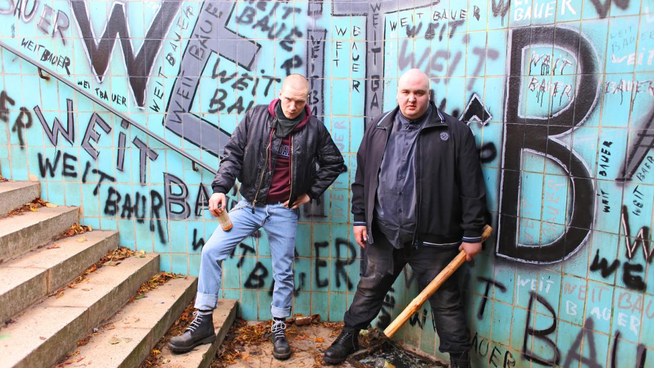 Heil, Szenenbild: Zwei Männer in schwarzer Kleidung stehen an einer mit Graffiti bedeckten Wand, einer hält eine Flasche, der andere einen Holzknüppel.