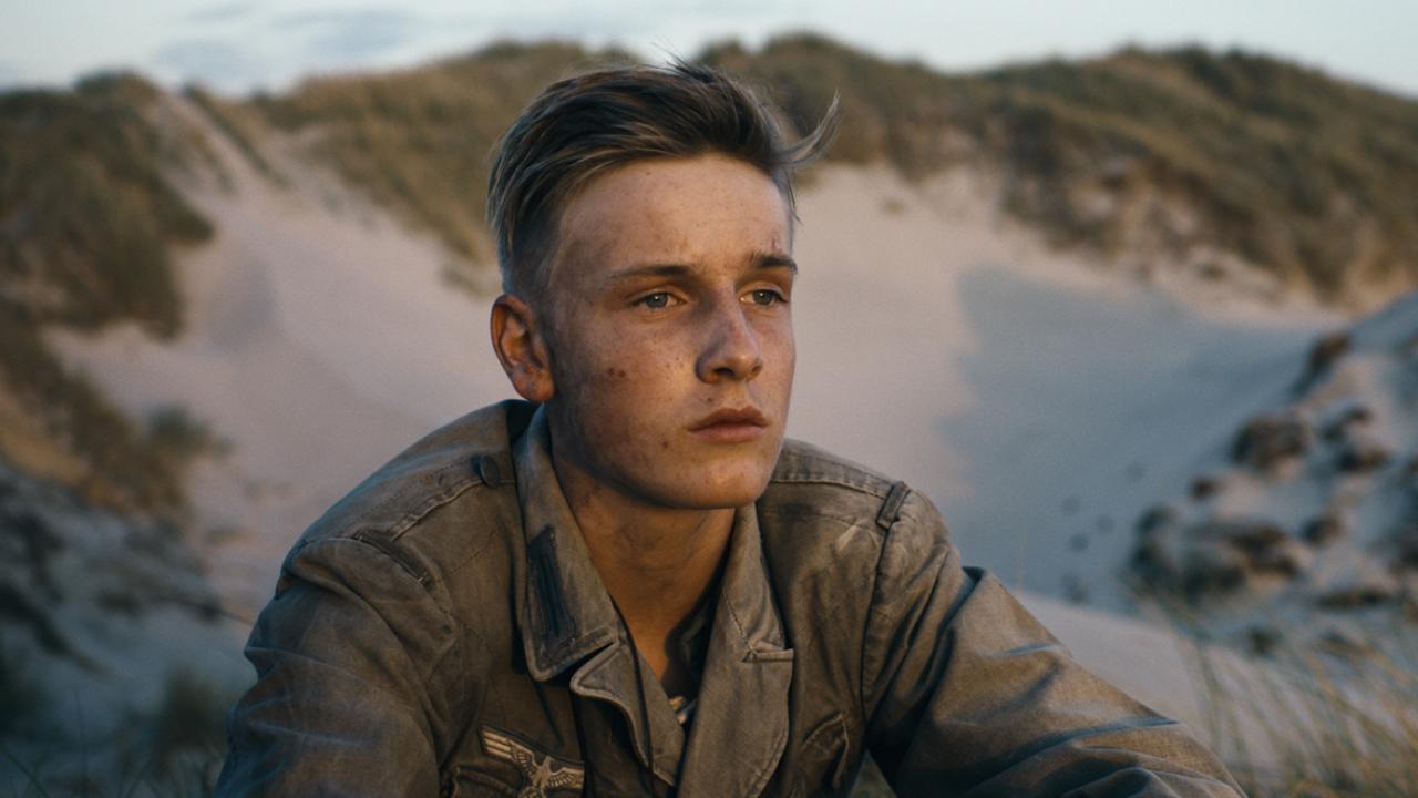 Unter dem Sand (Szenenbild): Ein junger Mann in Militärjacke und schmutigem Gesicht sitze zwischen Sanddünen und guckt traurig vor sich hin.