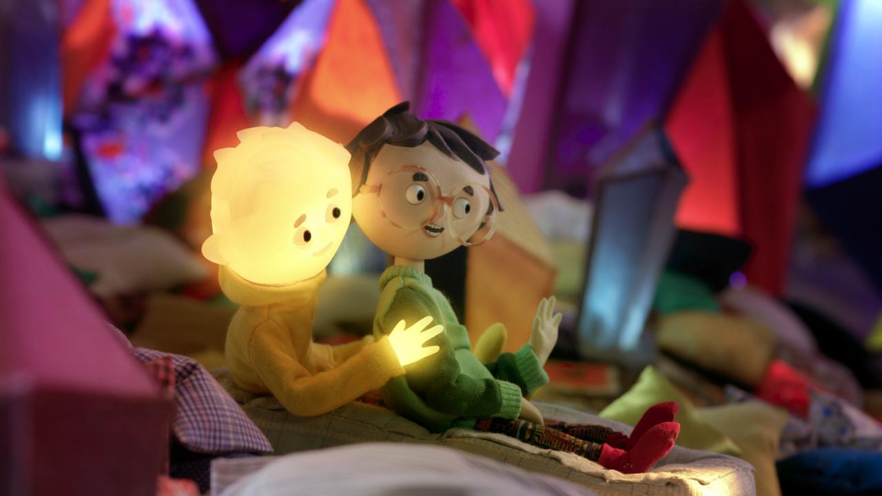 Szenenbild aus dem Puppentrickfilm TONY, SHELLY UND DAS MAGOISCHE LICHT: Zwei Kinder sitzen hintereinander und rutschen in einem farbenfrohen, höhlenartigen Raum nach unten. Der Kopf der Jungenfigur leuchtet.