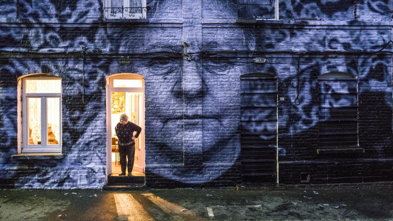 Augenblicke: Gesichter einer Reise, Szenenbild: Die Bewohnerin eines Straßenzugs im ehemaligen Bergbaugebiet Bruay-la-Buissière steht vor ihrem Haus. Auf der Hauswand ist ihr Porträt zu sehen.