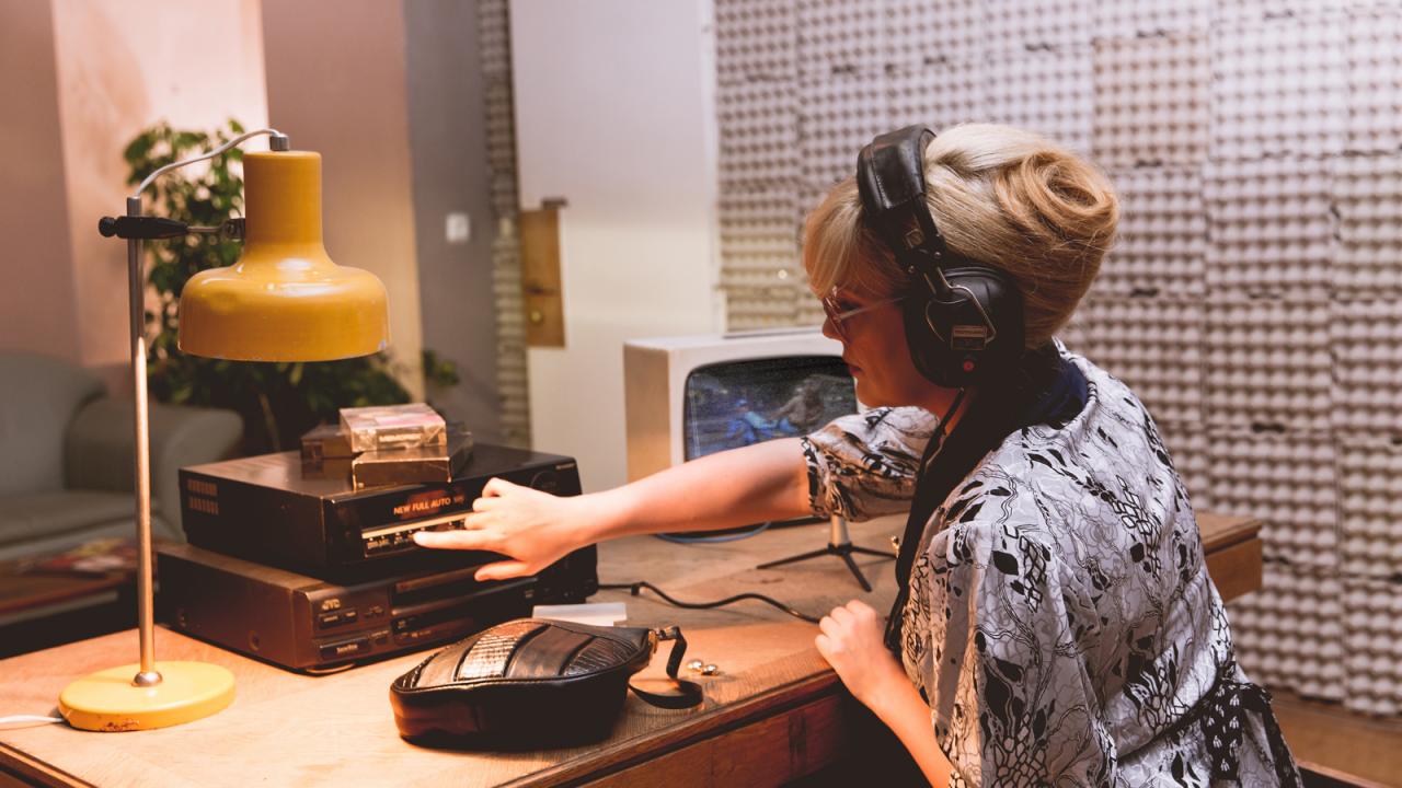 Chuck Norris und der Kommunismus, Szenenbild: Frau in Vintage-Kleidung und Kopfhörern bedient einen Videorekorder auf einem Retro-Schreibtisch mit gelber Lampe und altem Audio-Equipment.