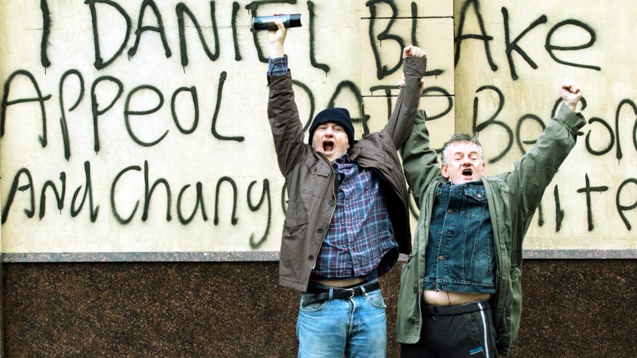 Szenenbild aus dem Drama ICH, DANIEL BLAKE: Zwei ältere Männer stehen mit hoch gestreckten Armen vor einer Mauer, auf die jemand "I Daniel Blake" gesprüht hat.