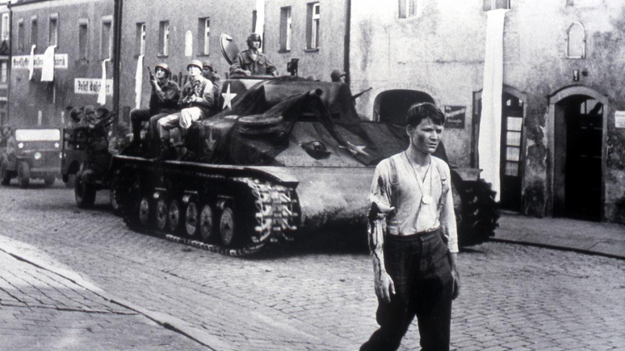 Szenenbild aus dem Drama DIE BRÜCKE: Ein junger Mann in Hemd und Uniformhose geht eine Straße entlang. Hinter ihm fährt ein Panzer.