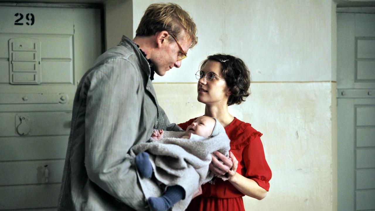 Szenenbild aus dem Drama IN LIEBE, EURE HILDE: Ein Mann und eine Frau, Hans und Hilde Coppi, stehen einander zugewandt in einem Gefängnisflur. Er hält ein Baby, ihren Sohn Hans, in den Armen.