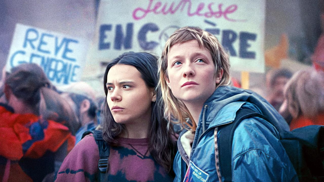 Szenenbild aus dem Coming-of-Age-Film TANDEM – IN WELCHER SPRACHE TRÄUMST DU?: Zwei junge Frauen stehen nebeneinander auf einer Demonstration in Frankreich. Sie schauen ernst. Im Hintergrund sind Menschen und Plakate zu sehen.