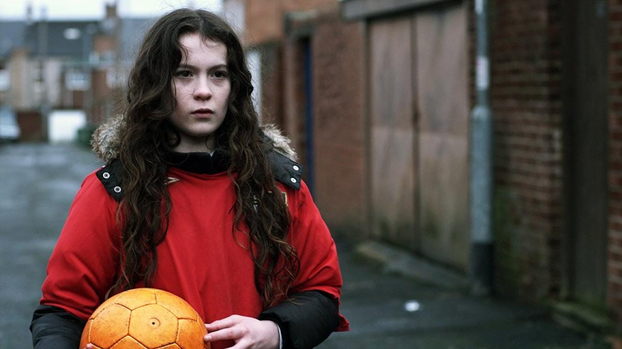 Der Ball, Szenenbild: Nahaufname eines dunkelhaarigen Mädchens in einem roten Pullover. Es steht in einer Gasse und hält einen Ball in den Händen.