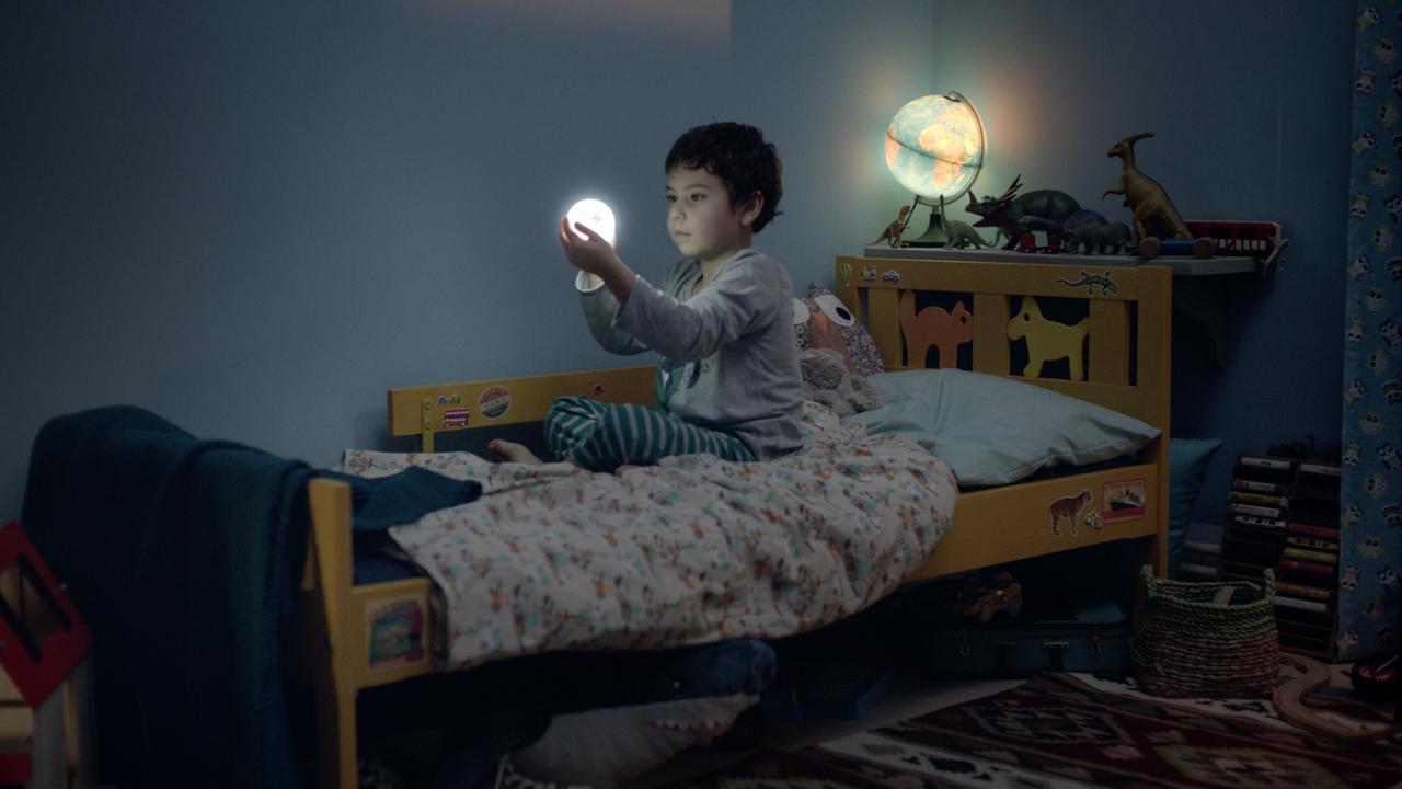 Der Mond und ich, Szenenbild: Ein kleiner Junge sitzt im Dunkeln in seinem Bett. Er hält eine kleine, strahlende Mondkugel in seinen Händen und betrachtet sie.