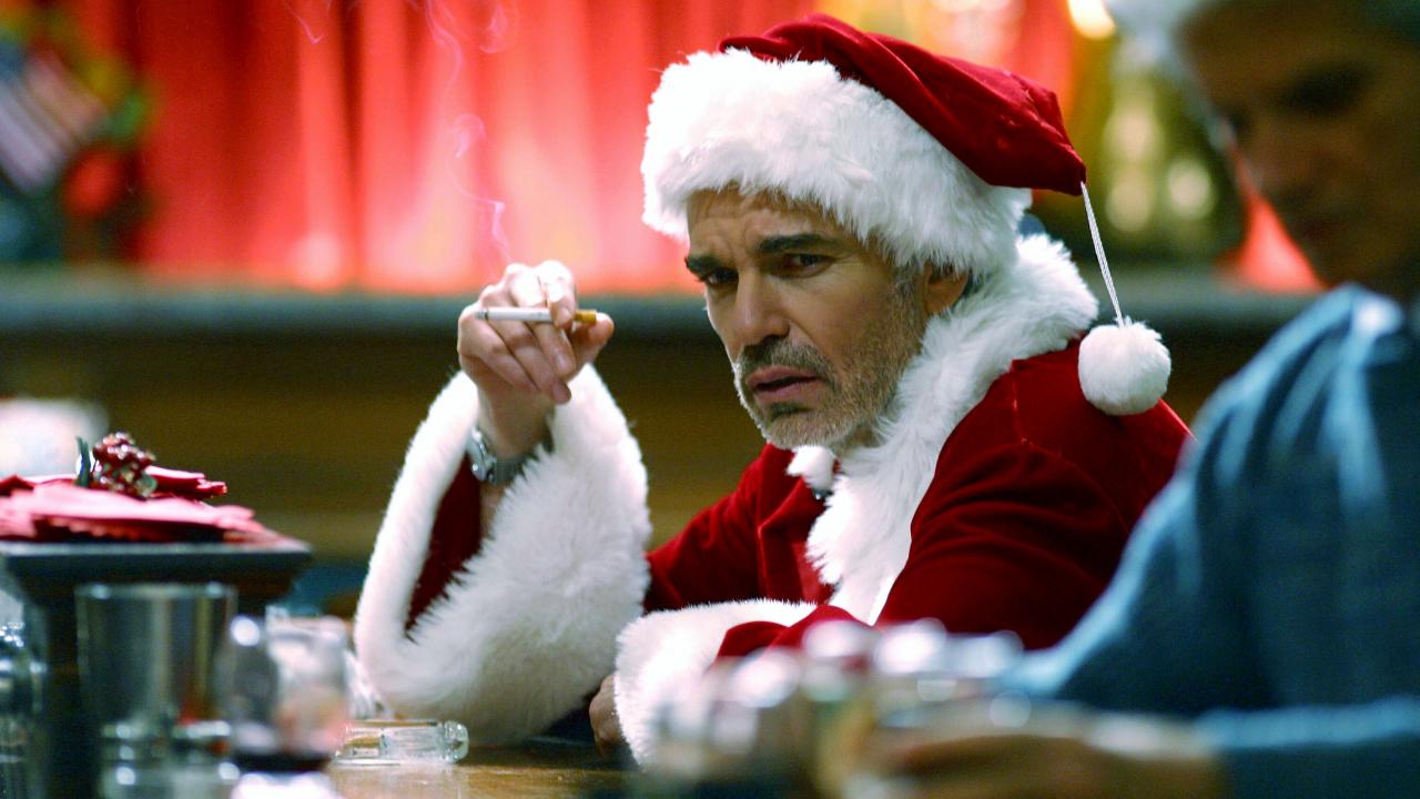 Filmstill aus BAD SANTA: Ein Mann im Weihnachtsmannkostüm sitzt rauchend an einer Bar und schaut grimmig in die Kamera.