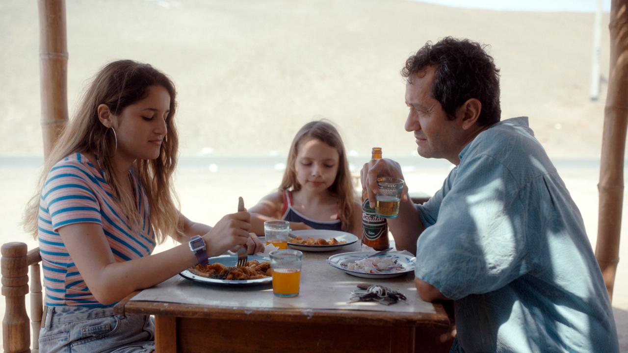 Szenenbild aus dem Drama REINAS – DIE KÖNIGINNEN: Zwei Mädchen im Teenageralter und ein Mann sitzen in einem Restaurant und essen. Im Hintergrund ist das Meer zu sehen.